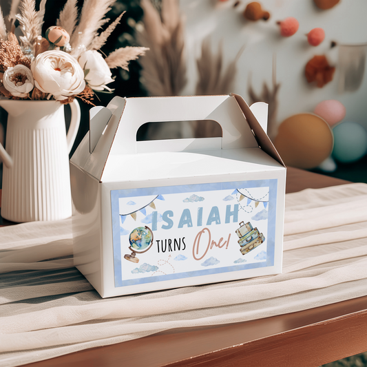 White gift box with 'Isaiah Turns One' label on a wooden surface with flowers and balloons in the background