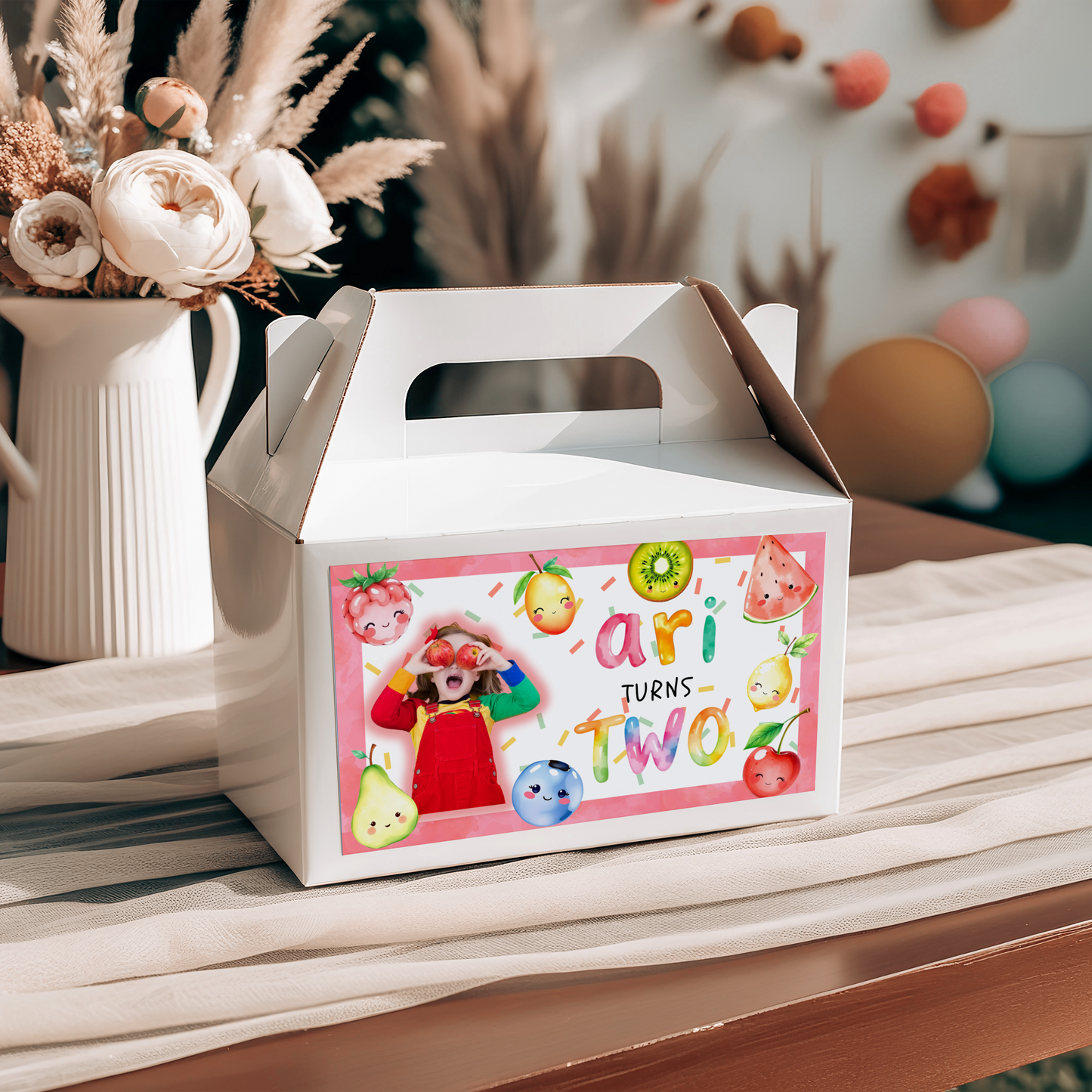 White party favor box with colorful design and text, placed on a table with flowers and balloons in the background.