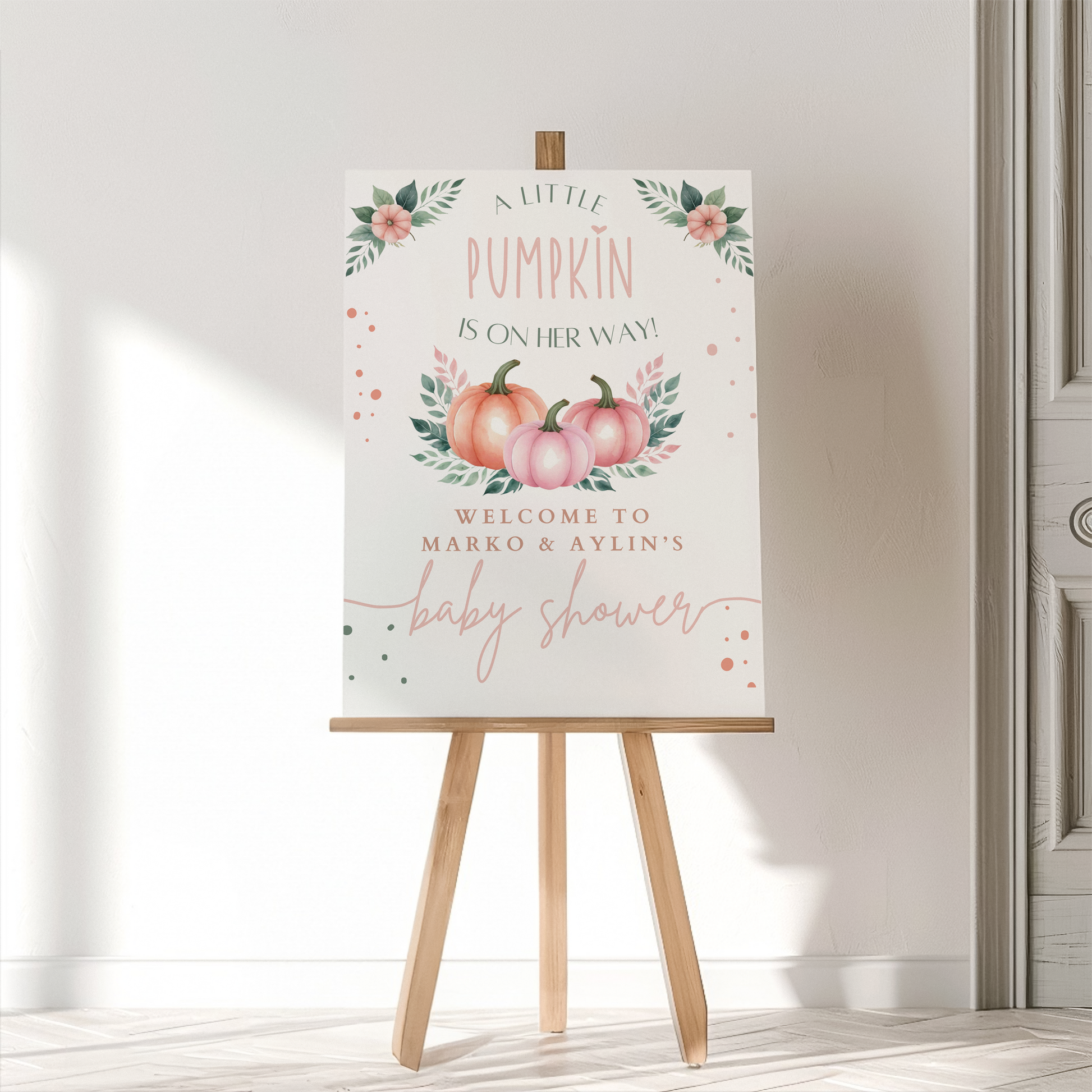 Baby shower sign with pumpkins and floral elements on an easel against a white wall.