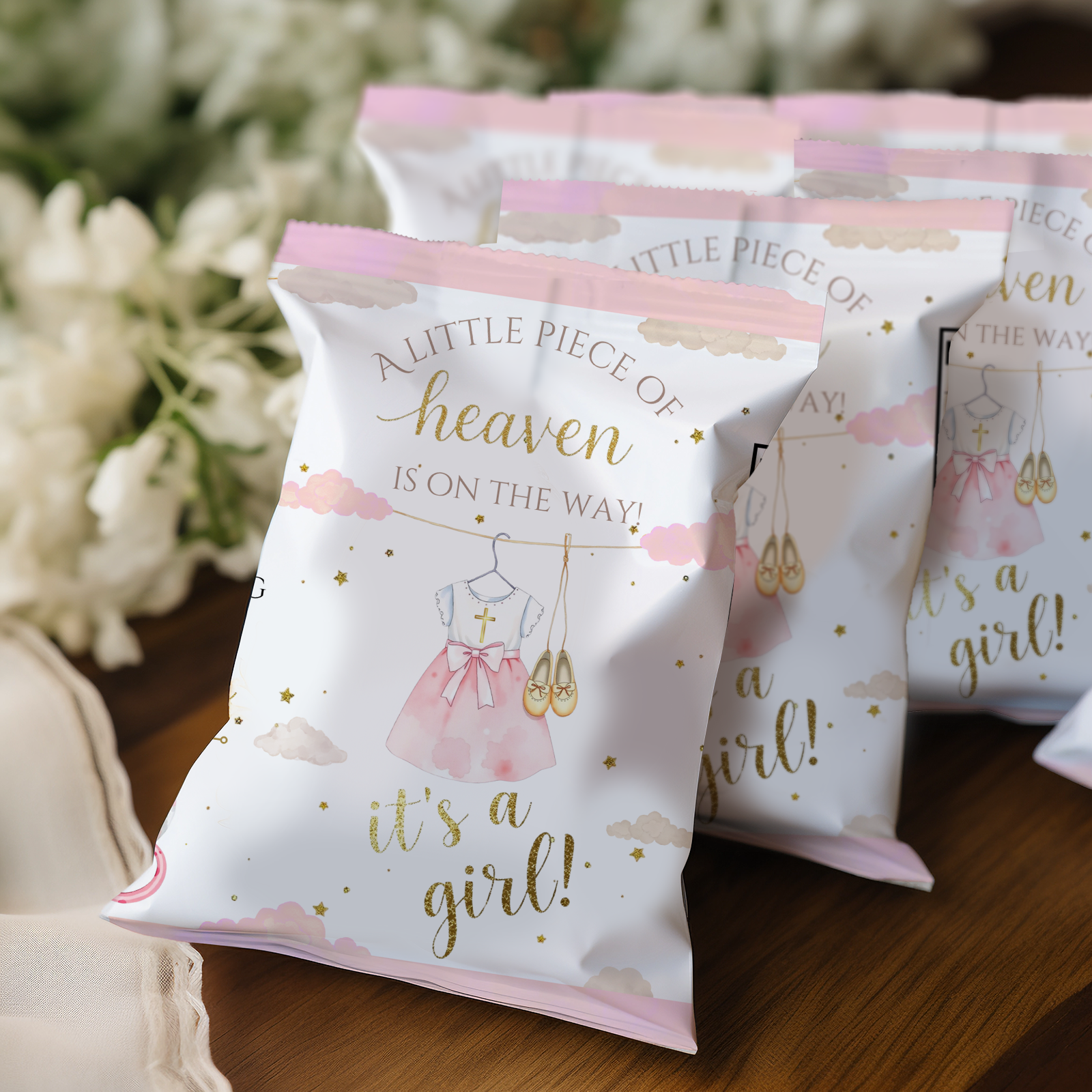 Baby shower favor bags with 'it's a girl!' text and pink dress illustration on a wooden surface.