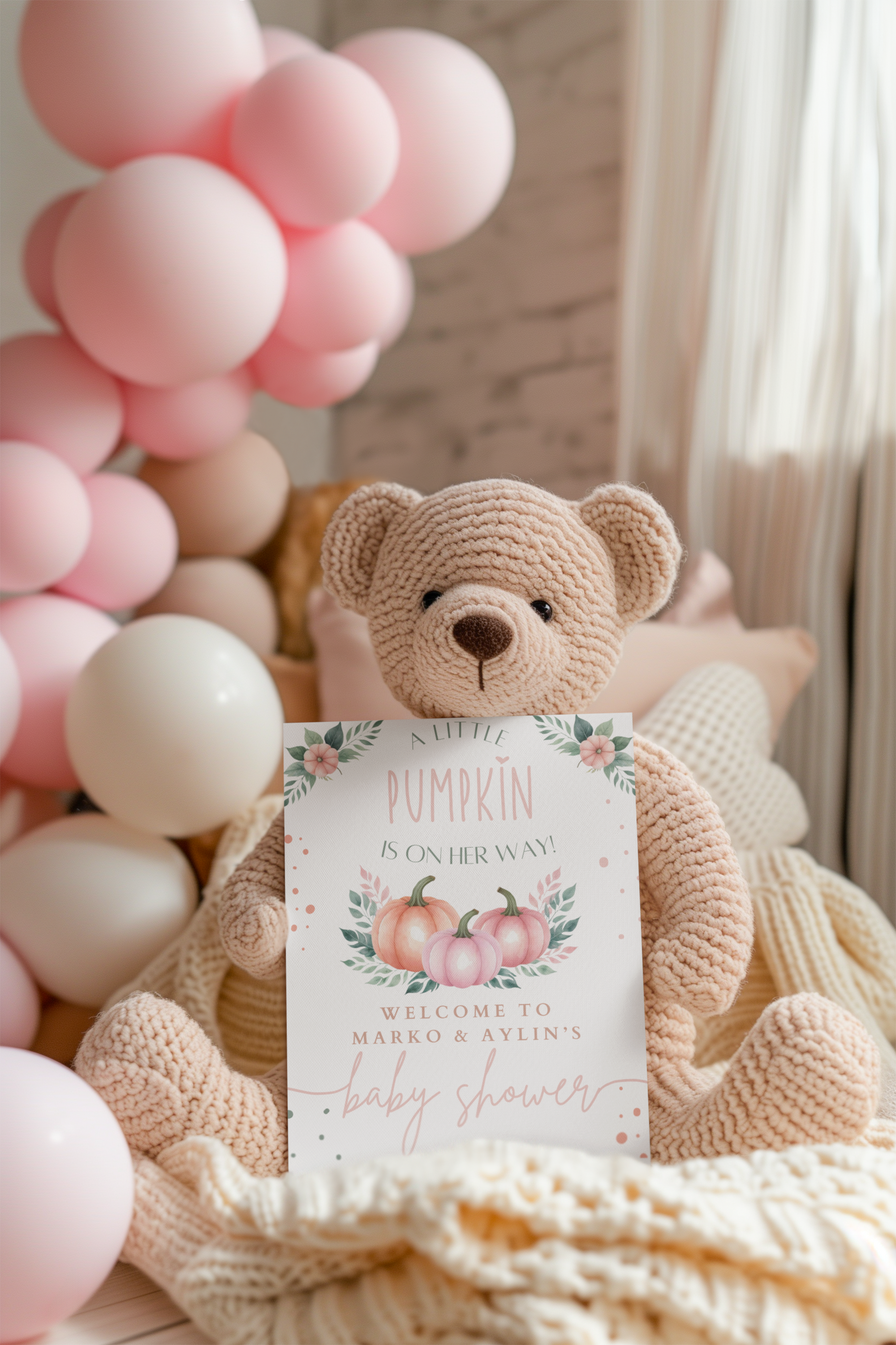 Crocheted teddy bear holding a baby shower invitation with pink balloons in the background