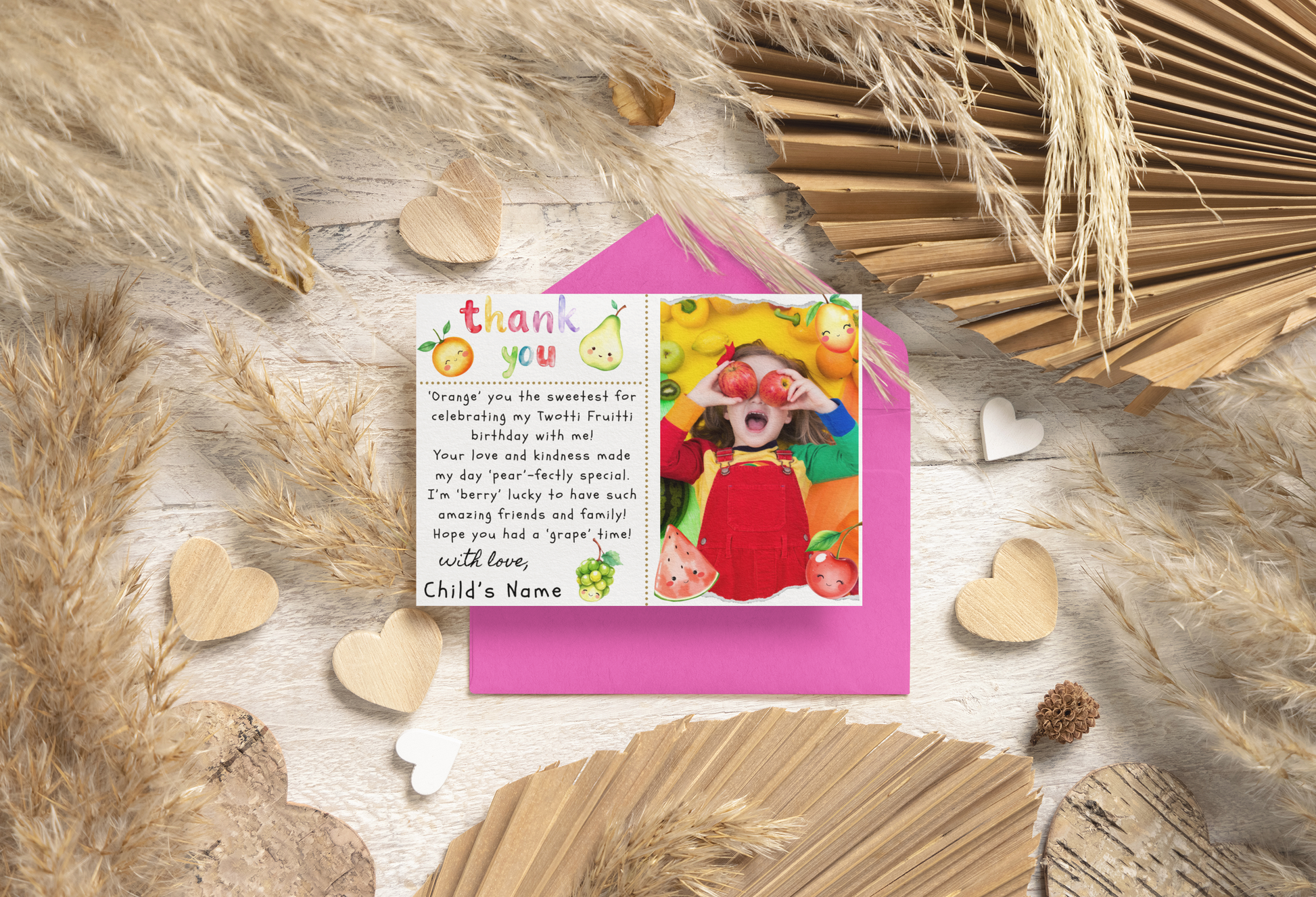 Greeting card with a photo of a child and text on a pink envelope, surrounded by decorative elements on a wooden surface.